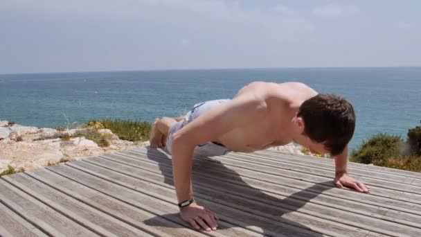 Athletic Caucasian man performs pushups on rocky coast beach on sunny day — Stock Video