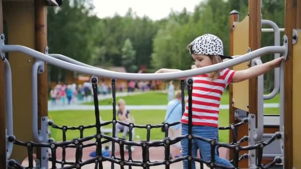 Una linda niña se mueve con éxito a través del puente de la red en el parque infantil . — Vídeos de Stock