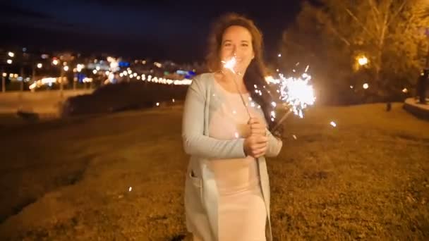 Smiling young woman stands on a high hill with sparkler with city night view on background — Stock Video