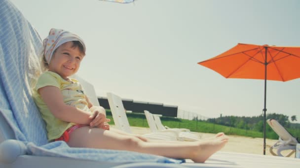 Glad liten baby flicka ta solbada i stol på stranden — Stockvideo