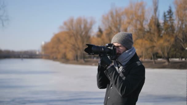 Νεαρός φωτογράφος με επαγγελματικό κάμερα εξοπλισμένη σε μαύρο μπουφάν λαμβάνοντας εικόνες από όμορφο χειμώνα πάρκο — Αρχείο Βίντεο