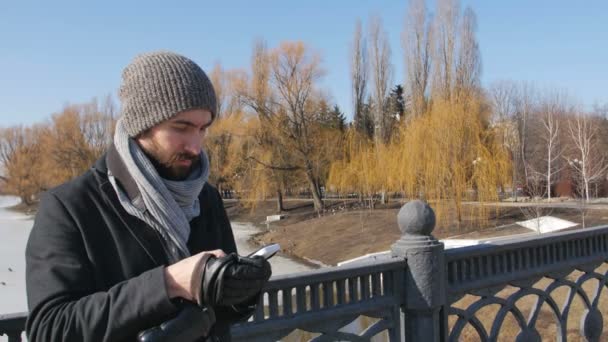 Человек в зимнем пальто использует телефон на фоне зимнего парка . — стоковое видео