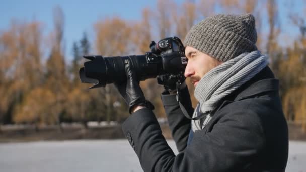 Młody mężczyzna, fotograf z profesjonalnym aparat wyposażony w czarna Parka biorąc Zdjęcia z pięknym Winter park — Wideo stockowe