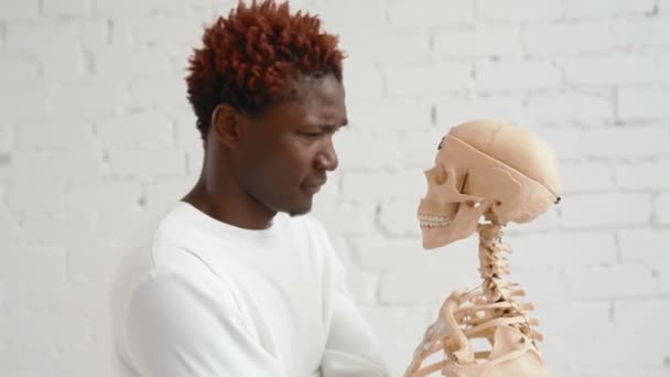 Безумный чёрный мужчина в смирительной рубашке танцует и смотрит на человеческий скелет — стоковое видео