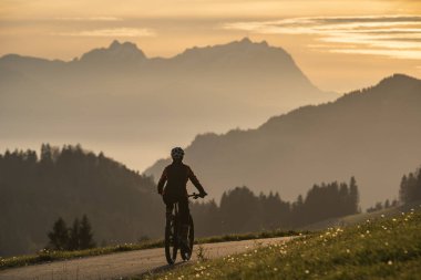 Aktif bir kadın gün batımında elektrikli dağ bisikletini sürüyor Saentis Dağı 'nın muhteşem siluetinin önünde, Appenzell Switthe