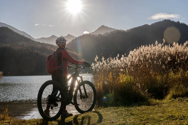 senior woman with electric mountain bike in bright backlit sun on the shore of lake Weissensee near city of Fuessen, eastern Allgaeu, Bavarian alps, Germany