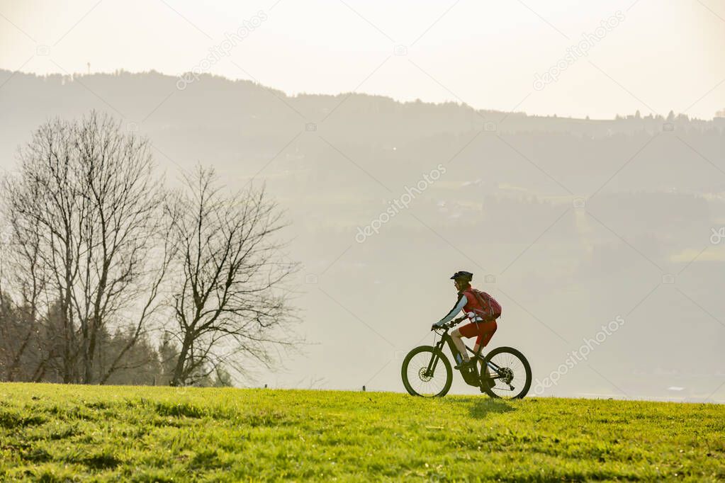 senior woman mountainbiking below the Nagelfluh mountain chain with Hochgrat summit on a e-mountainbike in early spring, in the Allgaeu Area near Steibis and Obers, a part of the bavarian alps,Germany