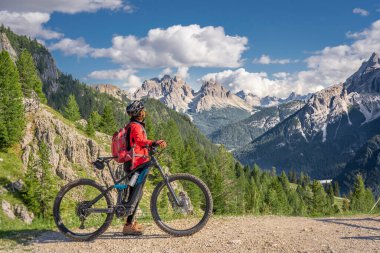 nice and active senior woman riding her electric mountain bike on the high plateau of Pratto Piazzo in the three peaks Dolomites , rocky silhouette of Mount Cristallo in background, South Tirol, Italy clipart