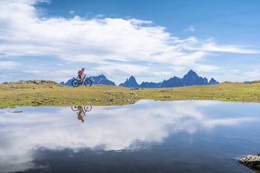 Üç Tepe Dolomitleri 'nde elektrikli dağ bisikletine binen hoş bir kadın kendini soğuk dağ gölünün mavi suyuna yansıtıyor.