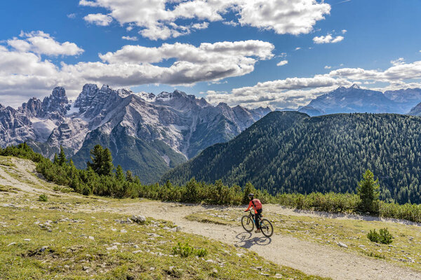 nice and active senior woman riding her electric mountain bike on the high plateau of Pratto Piazzo in the three peaks Dolomites , rocky silhouette of Mount Cristallo in background, South Tirol, Italy