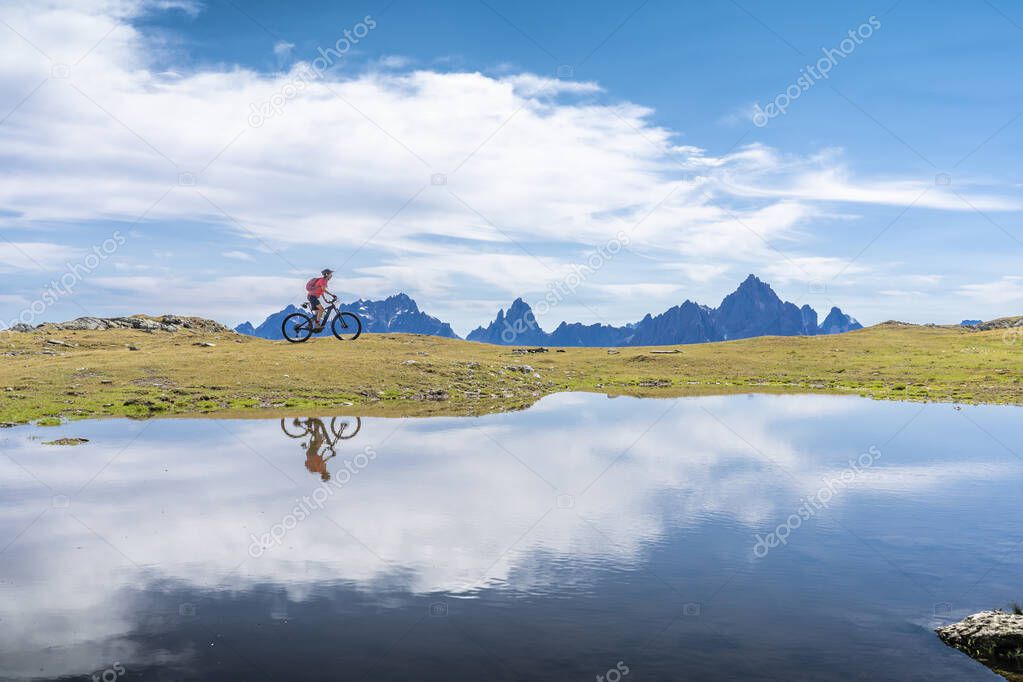 nice woman riding her electric mountain bike in the Three Peaks Dolomites, reflecting herself in the blue water of a cold mountain lake