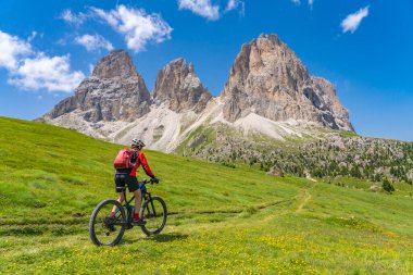 Selva Wolkenstein, Val Gardena, Güney Tirol ve Trentino, İtalya 'daki ünlü Sassolungo Dolomites zirvesinin altında elektrikli dağ bisikleti süren oldukça aktif yaşlı bir kadın.