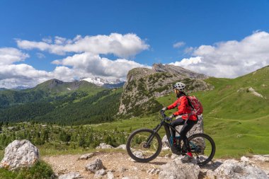 pretty active senior woman riding her electric mountain bike up to Valparola Pass in the Alta Badia Dolomites , South Tirol and Trentino, Italy clipart