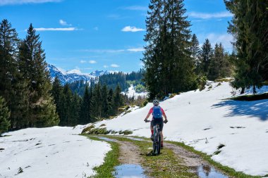 Güneşli bir baharın ilk günlerinde güneşli bir günde, arka planda yüksek Allgaeu dağları olan karların altındaki Gunzesried Vadisi 'nde elektrikli dağ bisikletine binen son sınıf güzeli bir kadın.