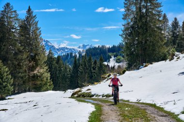 Güneşli bir baharın ilk günlerinde güneşli bir günde, arka planda yüksek Allgaeu dağları olan karların altındaki Gunzesried Vadisi 'nde elektrikli dağ bisikletine binen son sınıf güzeli bir kadın.