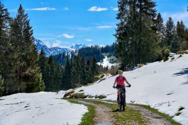 Güneşli bir baharın ilk günlerinde güneşli bir günde, arka planda yüksek Allgaeu dağları olan karların altındaki Gunzesried Vadisi 'nde elektrikli dağ bisikletine binen son sınıf güzeli bir kadın.