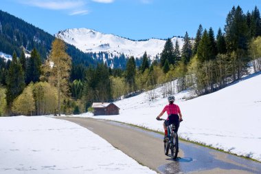 Güneşli bir baharın ilk günlerinde güneşli bir günde, arka planda yüksek Allgaeu dağları olan karların altındaki Gunzesried Vadisi 'nde elektrikli dağ bisikletine binen son sınıf güzeli bir kadın.
