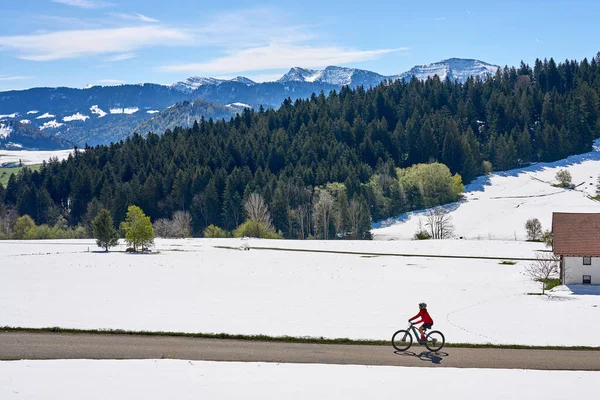 Allgaeu, Allgaeu, Oberstaufen yakınlarındaki Nagelfluh zincirinin kar kaplı dağlarının altındaki çayırlarda, güneşli bir baharın ilk günlerinde elektrikli dağ bisikletine binen yaşlı kadın. 