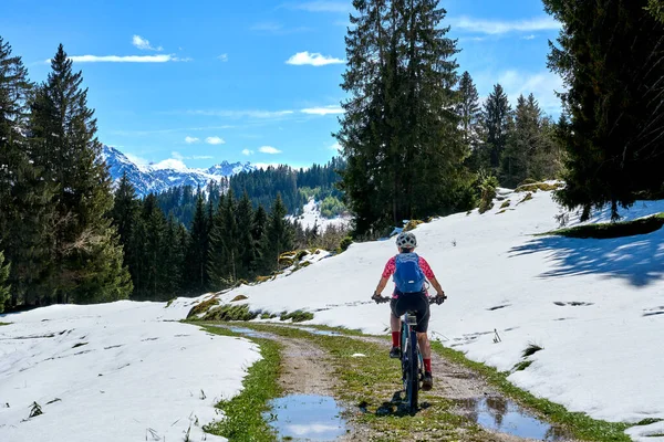 Güneşli bir baharın ilk günlerinde güneşli bir günde, arka planda yüksek Allgaeu dağları olan karların altındaki Gunzesried Vadisi 'nde elektrikli dağ bisikletine binen son sınıf güzeli bir kadın.