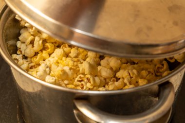Corn grains and a spices mass are poured into the kitchen pot to make popcorn. Popcorn can be seen along the narrow lid of the boiler clipart