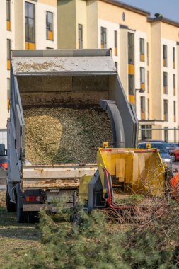 Güneşli bir bahar çalışma gününde, ağaç kesicisinin çöp kamyonu, bir çim biçme makinesiyle birlikte arka planda evleri olan dolu bir çayırda bulunur..