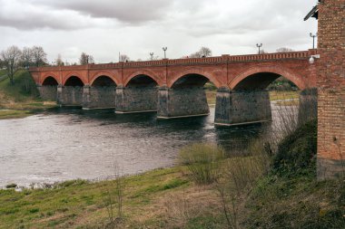 Venta Nehri 'nin bahar manzarası ve gri bulutlu gökyüzü ile Eski Kuldiga Tuğla Köprüsü.