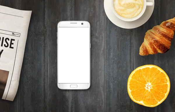 Smart phone with isolated display for mockup on table. Newspapers, coffee, croissant and orange on table. Top view.
