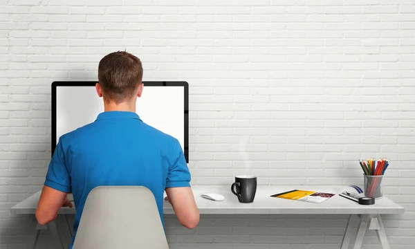 Muž pracující na počítači s izolovanou obrazovkou ve vnitrozemí. Pracovní stůl s klávesnicí, myší, šálkem kávy, papírem, tužkami. Volné místo na zdi pro text. — Stock fotografie