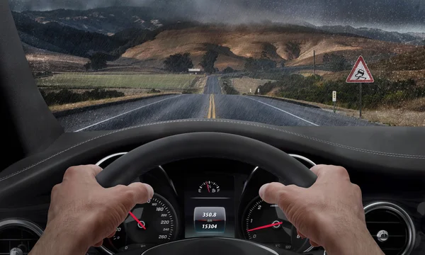 Rijden in de regen. Uitzicht vanaf de hoek van de bestuurder terwijl de handen op het wiel. Is een teken voor gladde weg langs de weg. Regen gevlekte voorruit. — Stockfoto