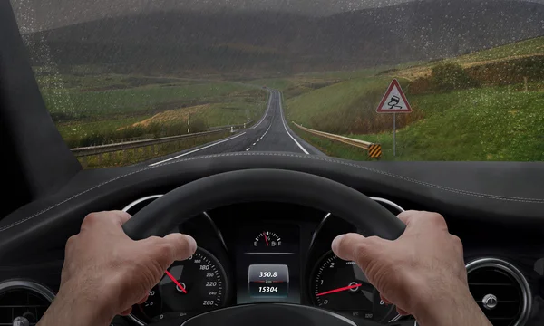 Conduciendo con tiempo lluvioso. Vista desde el ángulo del conductor mientras las manos en el volante. Junto a la carretera hay una señal de camino resbaladizo. Lluvia salpicada parabrisas . —  Fotos de Stock