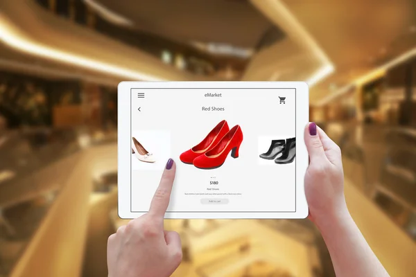 Woman online shopping with tablet. Holding device and choose red shoes. Shopping center in the background. — Stock Photo, Image