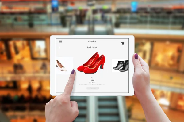 Woman online shopping with tablet. Holding device and choose red shoes. Shopping center in the background. — Stock Photo, Image