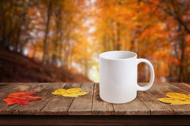 Renkli sonbahar yaprakları ve bulanık orman arkaplanı olan kırsal masada beyaz kahve fincanı ürün tasarım modelleri için idealdir.