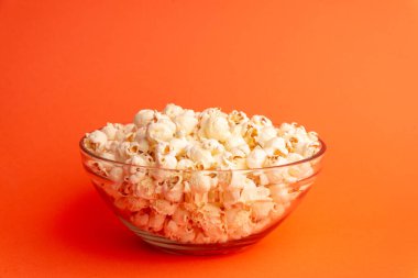 Patlamış mısırın turuncu arka plandaki cam kasenin üzerine düşmesiyle dinamik stüdyo görüntüsü, eğlence, hareket ve neşeli atıştırma anlarını simgeliyor.