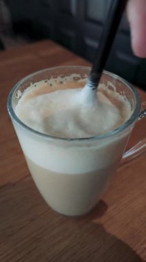 Yatay yakın çekim, cam bardakta köpüklü latte kahve, ahşap masa, samimi ve davetkar kafe atmosferi.