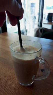 Elle kahveyi samanla karıştıran dikey video, modern kafe dekorasyonu, doğal ışık ve yaşam tarzı görselleri için yumuşak rahat renk tonları