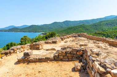 Ancient stone ruins positioned above a turquoise sea and green coastal hills, captured in bright daylight, showcasing historic architecture within a scenic Mediterranean landscape