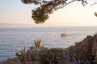 Sakin denizdeki geleneksel yelkenli teknesi kıyı bitkileri ve sıcak akşam ışığı ile kayalık sahilden izleniyor.