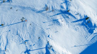 Kayakçılar, uzun gölgeler ve dokulu kar yüzeyli alp kayak yamaçlarının yukarıdan görünüşü kış sporları aktivitelerini ve tatil köylerinin altyapısını gösteriyor.