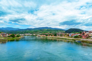 Bosna-Hersek 'in Višegrad kentinden akan Drina Nehri' nin geniş manzarası, kasaba binaları, yeşil tepeler, dramatik gökyüzü ve huzurlu yaz atmosferiyle çevrili..