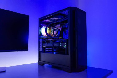 Masaüstü bilgisayarı rgb soğutma vantilatörü, sıvı soğutmalı işlemci ve grafik kartı ile oynatılıyor, koyu modern çalışma alanında monitörün yanına yerleştirilmiş.