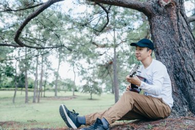 Asyalı adam çam ağacının altında gitar çalıp şarkı söylüyor..