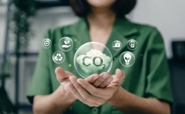 Karbon azaltımını, yeşil enerjiyi, geri dönüşümü ve sürdürülebilir çevresel çözümleri temsil eden yüzen bir CO2 bulut simgesini tutan eller.