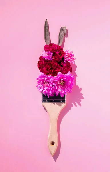 2021 Paskalya kompozisyonu. Pembe, kırmızı, mor çiçeklerden, boya fırçasından ve pastel arka planda tavşan kulaklarından oluşan yaratıcı bir düzen. Düz yatış, üst görünüm.