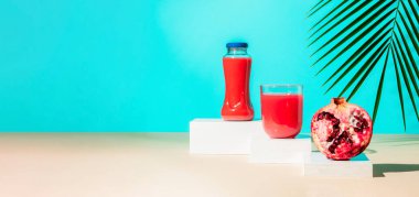 Yeni kesilmiş nar meyvesi ve podyumlarda meyve suyu ya da tropikal palmiye yaprağıyla mavi arka planda kaidesi olan modern natürmort yaşam. Minimum detoks diyeti ya da yaz konsepti. Boşluğu kopyala.