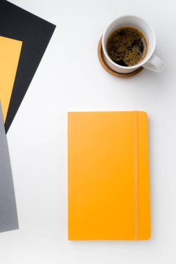 Beyaz masa yüzeyi, sağ üst köşesinde beyaz kahve fincanı ve ahşap sıcak standı var. Sağ alt köşede sarı defter. üç yaprak kağıt gri sarı ve siyah A4 biçimi
