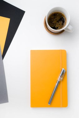 Beyaz masa yüzeyi, sağ üst köşesinde beyaz kahve fincanı ve ahşap sıcak standı var. Sağ alt köşede üzerinde gri kalem olan sarı defter. Üç yaprak kağıt grisi sarı