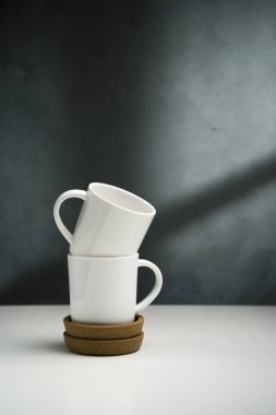 Arka plan koyu beton duvar ve pencereden yansıyan ışık. Beyaz masa. Çerçevenin ortasındaki masada, iki beyaz seramik parlak bardak bir diğerinin üstüne istiflenir. Mantar