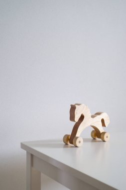 Beyaz bir masanın arka planında beyaz bir duvar köşesine karşı. Sağ alt köşedeki masada tekerlek üzerinde kahverengi tahta bir at var. Metin için boşluk. Pencereden sağa doğru ışık