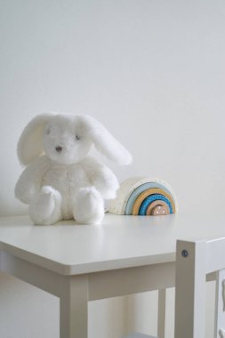 Beyaz bir duvarın arkasında, beyaz bir masanın köşesinde ve beyaz bir sandalyede. Sol alt köşedeki masada yumuşak, tüylü bir tavşan oturuyor. Masanın üzerinde oyuncak gökkuşağı mavisi bej rengi